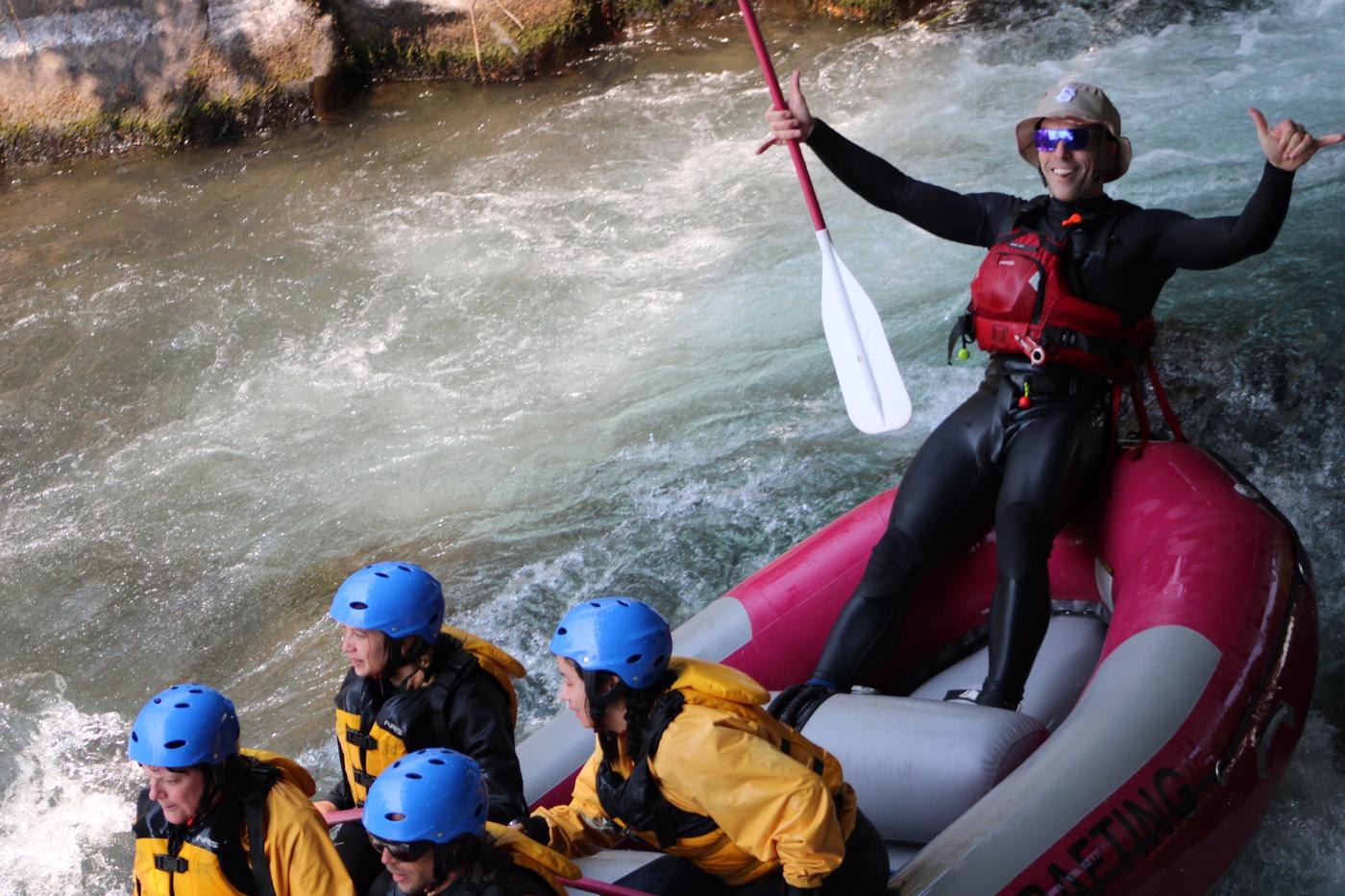 Standing on the tube of a raft mid-river, paddle raised, throwing a shaka